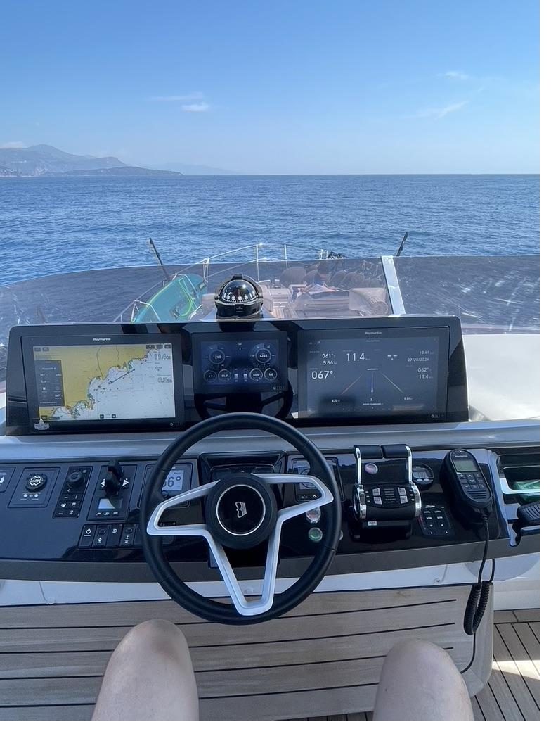 Flybridge helm station of a motor yacht underway — wheel, chartplotter and instrument displays, the Côte d'Azur coastline ahead